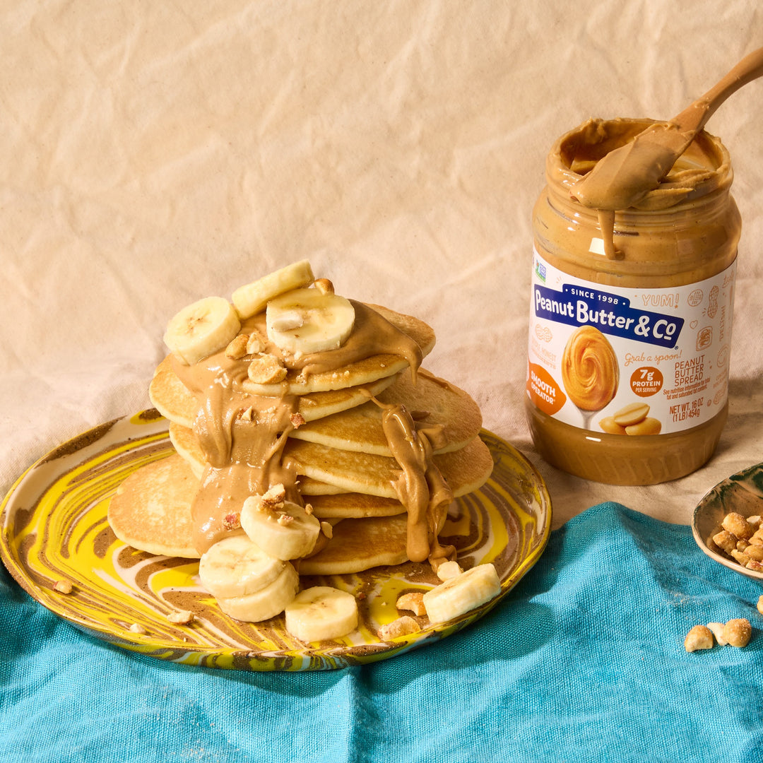 Stack of pancakes with peanut butter and banana slices on a plate next to a jar of Peanut Butter & Co. peanut butter.