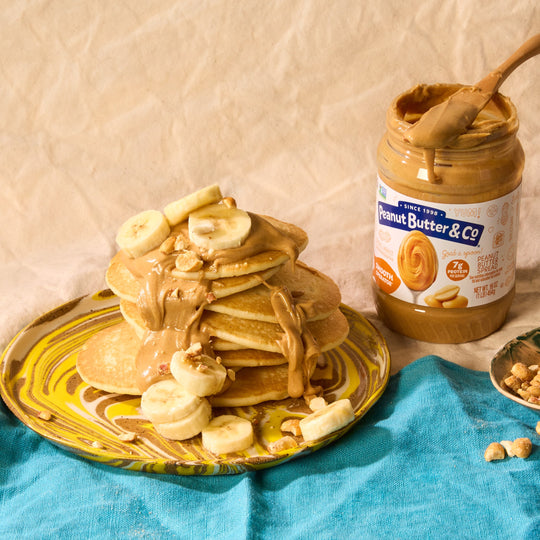 Stack of pancakes with peanut butter and banana slices on a plate next to a jar of Peanut Butter & Co. peanut butter.